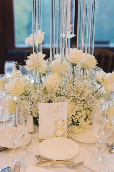 Reception tablescape with a wedding table centerpiece of white roses and baby’s breath in tall glass cylinder vases, gold table number 8 by a window