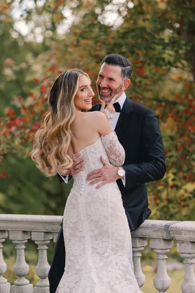 Couple portrait of bride and groom embrace, bride looking over shoulder in lace wedding dress by stone balustrade with autumn trees behind