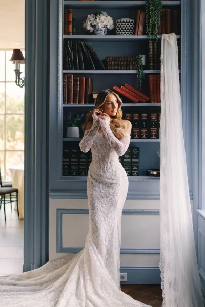 Bridal portrait in a lace wedding dress with a long veil and train, standing by a blue library bookshelf in soft window light