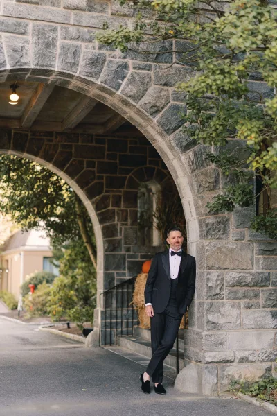 Groom portrait in a black tuxedo and bow tie, leaning by a stone archway on an outdoor walkway with greenery and steps behind him