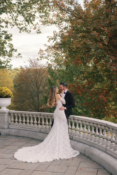 Couple portrait of bride and groom embrace, bride touching his face on a stone terrace with balustrade and autumn trees under cloudy sky