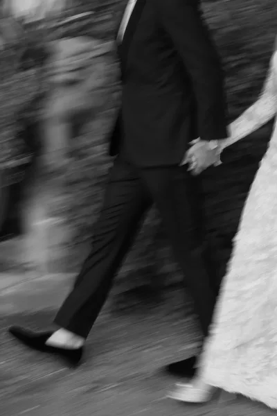 Wedding couple walking hand in hand, groom in black tuxedo beside bride in lace wedding dress along an outdoor garden pathway by a stone wall