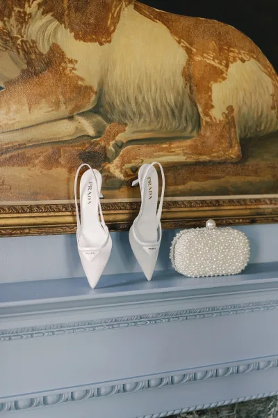 Bridal shoes in a wedding shoes flatlay with white slingback heels and a pearl clutch on a mantel shelf against a blue wall and framed painting