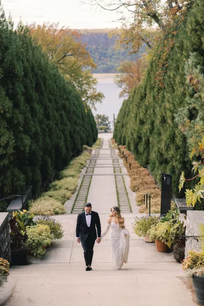 Couple portrait of bride and groom walking hand in hand on stone stairs, her veil flowing beside potted plants on a tree-lined path