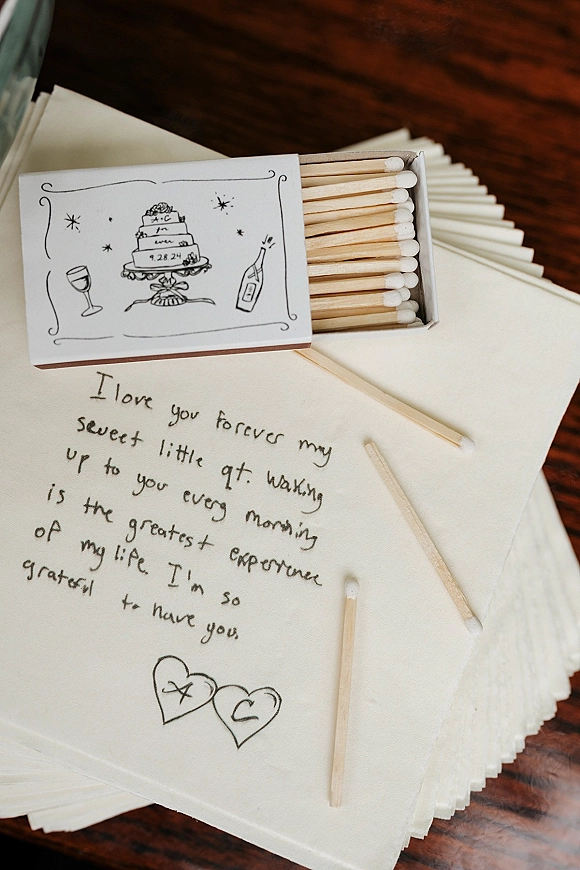 Wedding matchbooks and custom matchboxes wedding favor arranged with cocktail napkins, matches, and a handwritten note on a wood table flat lay