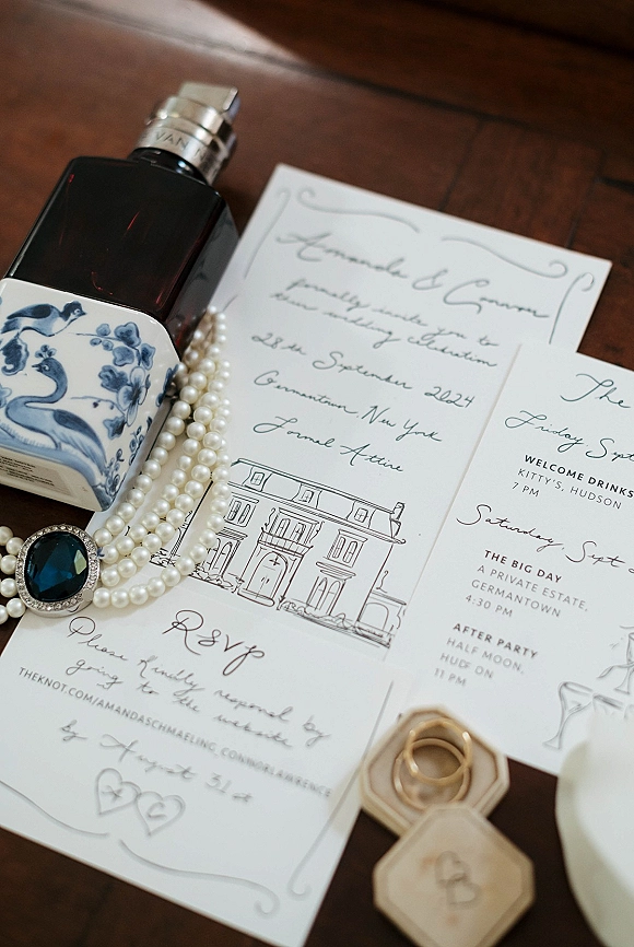 Invitation suite with calligraphy wedding invitation, RSVP and details cards, perfume, pearl necklace and rings arranged on a wood table flat lay