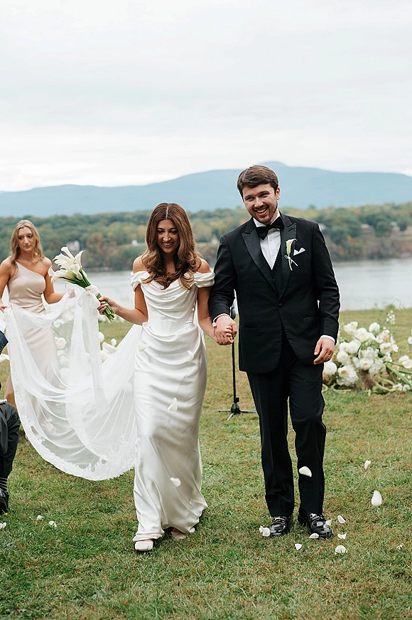Wedding recessional as bride and groom walk hand in hand through rose petals, her long veil and calla lily bouquet by a lakeside mountain lawn