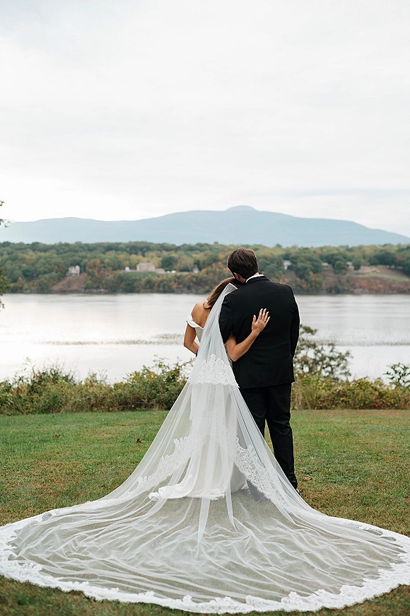 Couple portrait of bride and groom from behind, arm in arm with lace veil train, overlooking a mountain lake under cloudy skies