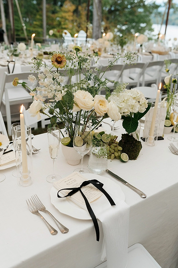 Reception tablescape with taper candles, white rose and hydrangea centerpiece, black ribbon place setting, and fruit on a garden table by a lake