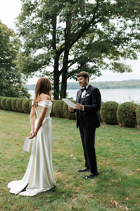 Wedding vows during an outdoor vow exchange as groom reads vow cards beside bride in an off-the-shoulder gown on a lakeside lawn