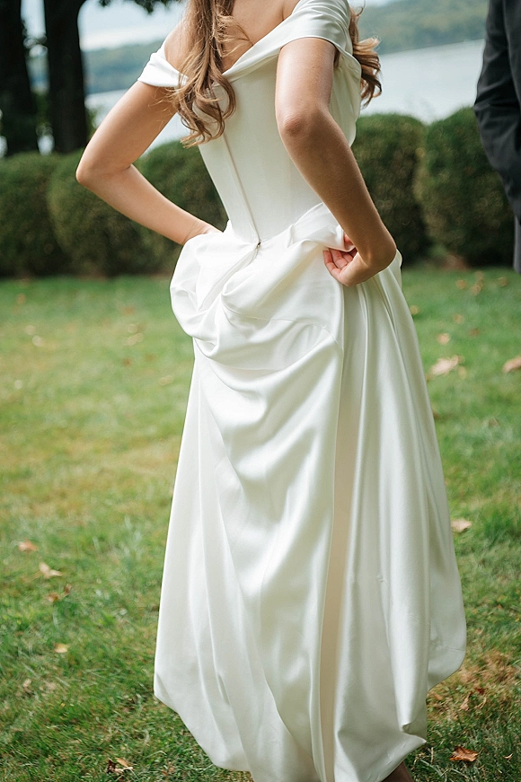 Wedding dress with an off the shoulder neckline in satin, showing a draped skirt and back zipper on a lawn by a lake and hills