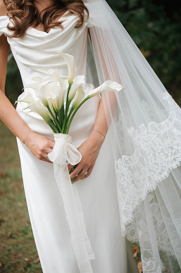 Bridal portrait of a bride holding bouquet of white calla lilies, showing her satin dress, lace veil, and ring against greenery