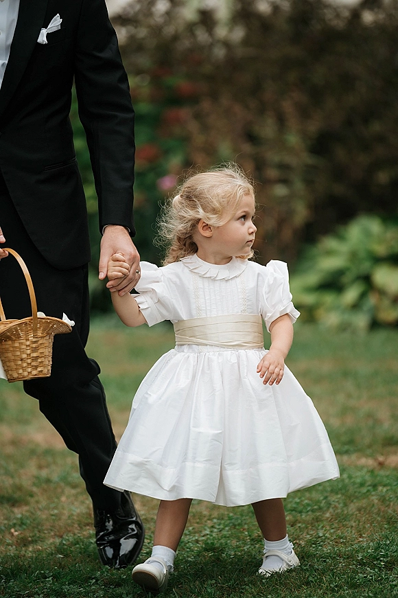 Flower girl in a flower girl dress with puff sleeves and satin sash holds a wicker basket beside a tuxedoed child on a garden lawn