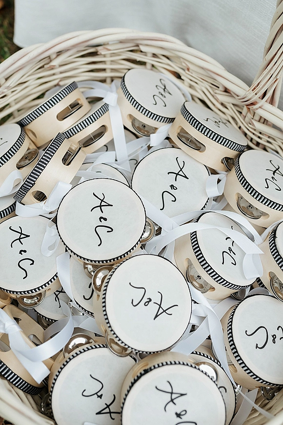 Wedding favor tambourines with personalized calligraphy, white ribbon, and black striped trim displayed in a wicker basket on grass