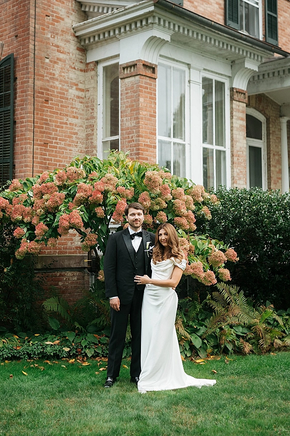 Couple portrait of bride and groom posing on a lawn, her off-the-shoulder satin dress and his tuxedo, by a brick house and hydrangeas