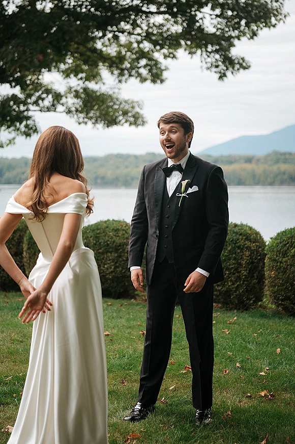 Wedding first look as bride in an off the shoulder dress approaches groom in black tuxedo on a lakeside lawn with mountains under clouds