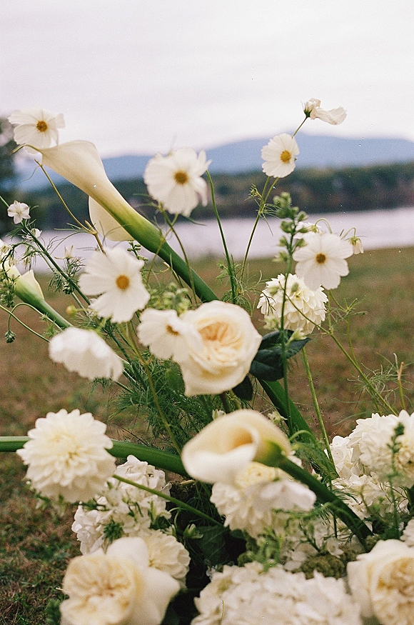 Wedding florals with white wedding flowers, including calla lilies, daisies, roses, and greenery arranged on grass by a lake and hills