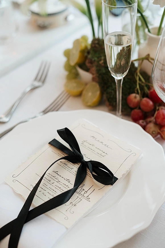 Wedding place setting with a wedding menu card tied in black ribbon on a plate, surrounded by glassware, silverware, candles, and fruit on a white tablecloth