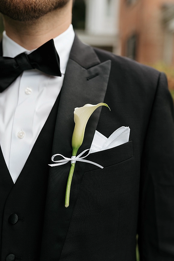 Groom tuxedo with black tie groom details, black bow tie and calla lily boutonniere on satin lapel, greenery behind