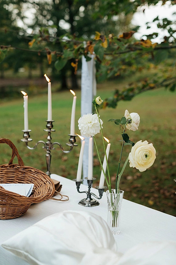 Wedding tablescape with candelabra wedding centerpiece, white taper candles and blooms on a white tablecloth set on a garden lawn under trees