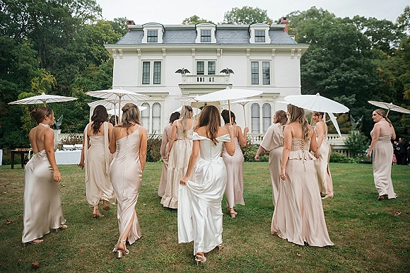 Bridesmaids walking in champagne bridesmaid dresses, carrying white parasols on a mansion lawn with trees and outdoor reception tables