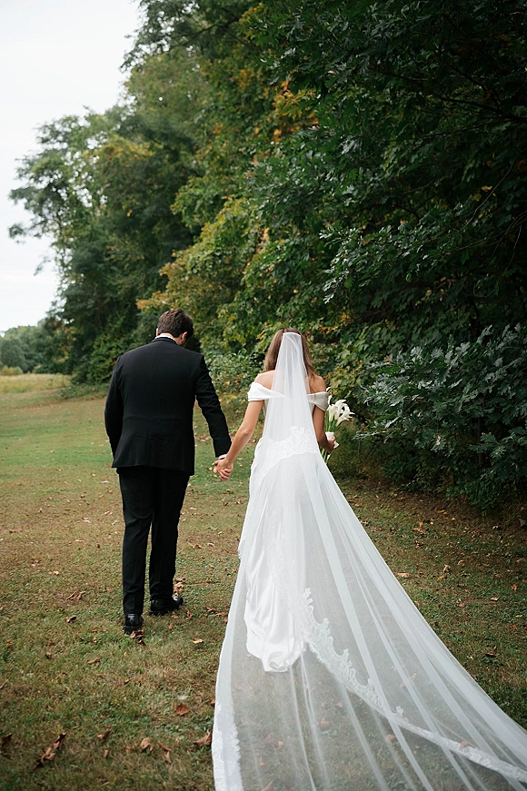 Couple portrait of bride and groom walking away holding hands, her long veil trailing, bouquet in hand, across a grassy field with trees