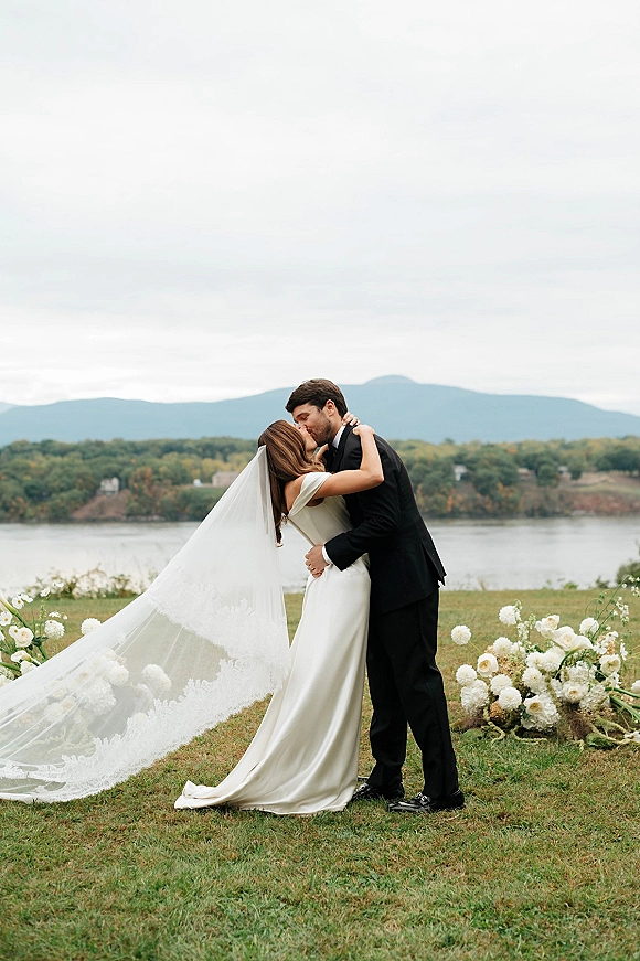 Wedding kiss portrait of bride and groom kissing, long veil trailing as he holds her waist on a lakefront lawn with mountains under clouds