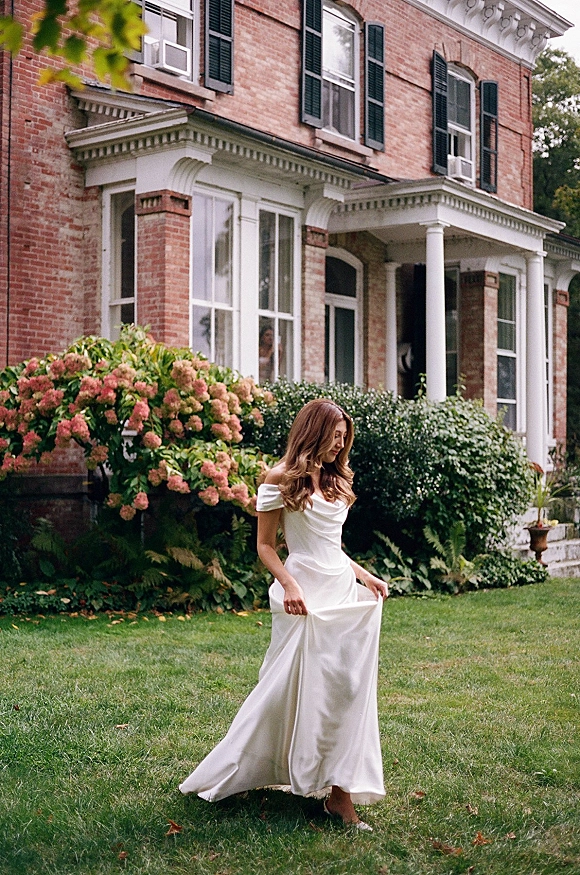 Bridal portrait of a bride in an off the shoulder wedding dress, holding her hem on a lawn before a brick manor with white columns