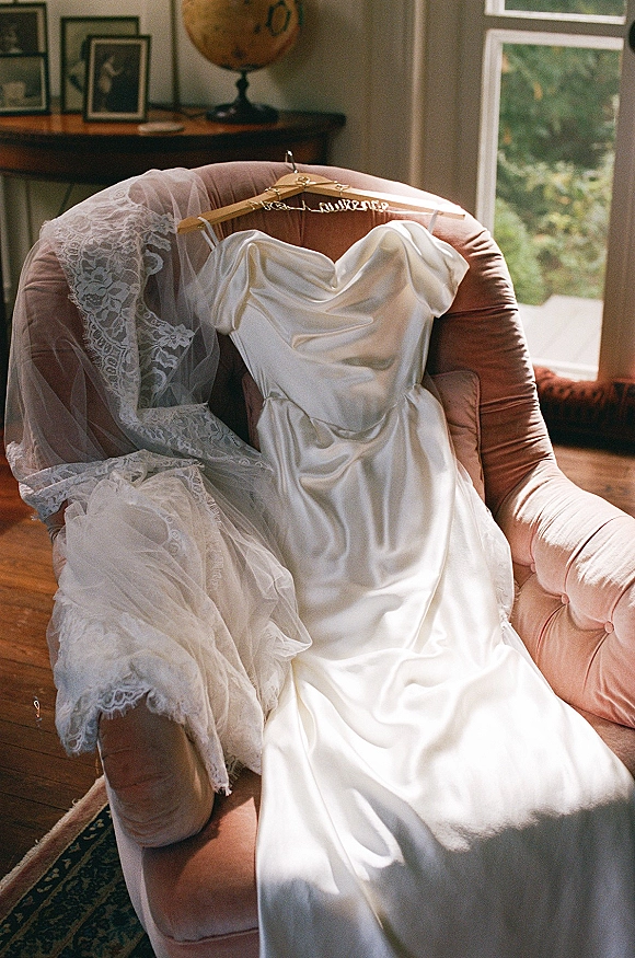 Wedding dress in silk satin draped over an upholstered armchair with a lace veil, beside a window and hardwood floor in natural light