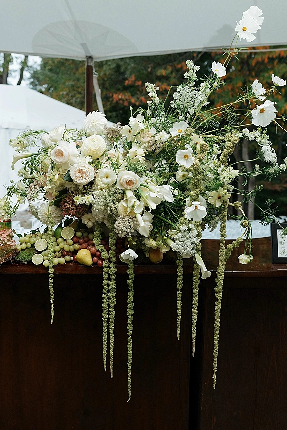 Wedding floral centerpiece with white roses and cosmos, hanging amaranthus and fruit on a rustic wood table under a tent canopy