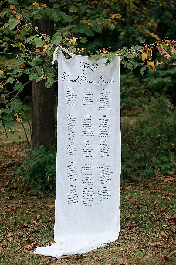 Wedding seating chart on a linen seating chart banner with ribbon ties and calligraphy, hanging from tree branches over grass and leaves