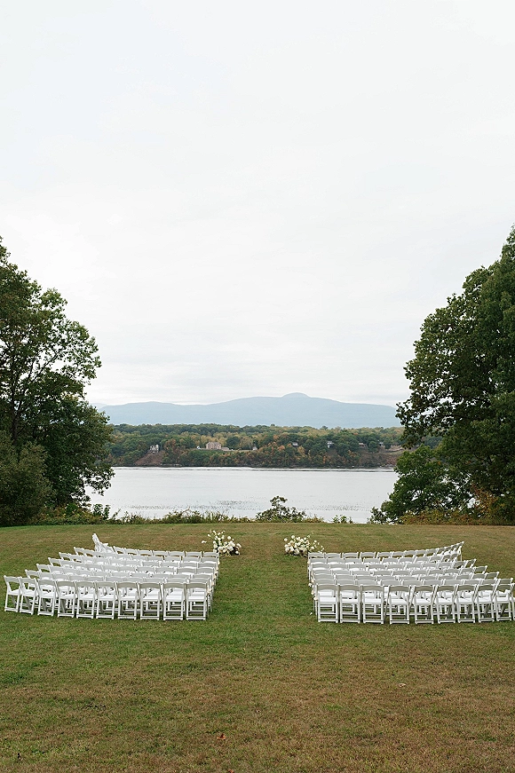 Ceremony setup with white folding chairs in rows and aisle florals on a grass lawn, facing a lake with trees and distant hills