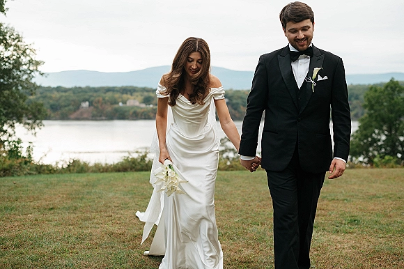 Couple portrait of bride and groom walking hand in hand, her veil and bouquet flowing on a lakeside lawn with mountains behind