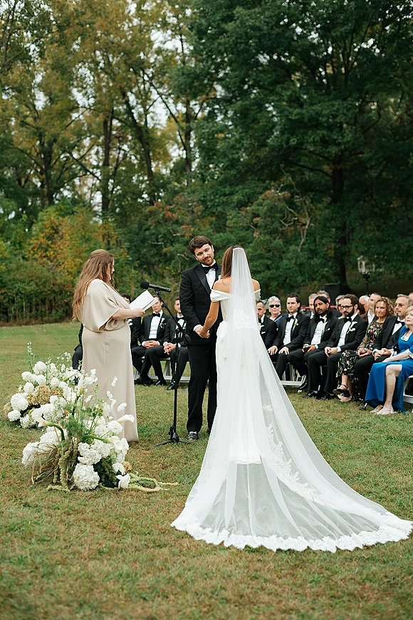 Wedding vows as bride and groom hold hands at an outdoor wedding ceremony, her cathedral veil and lace train on a garden lawn with guests