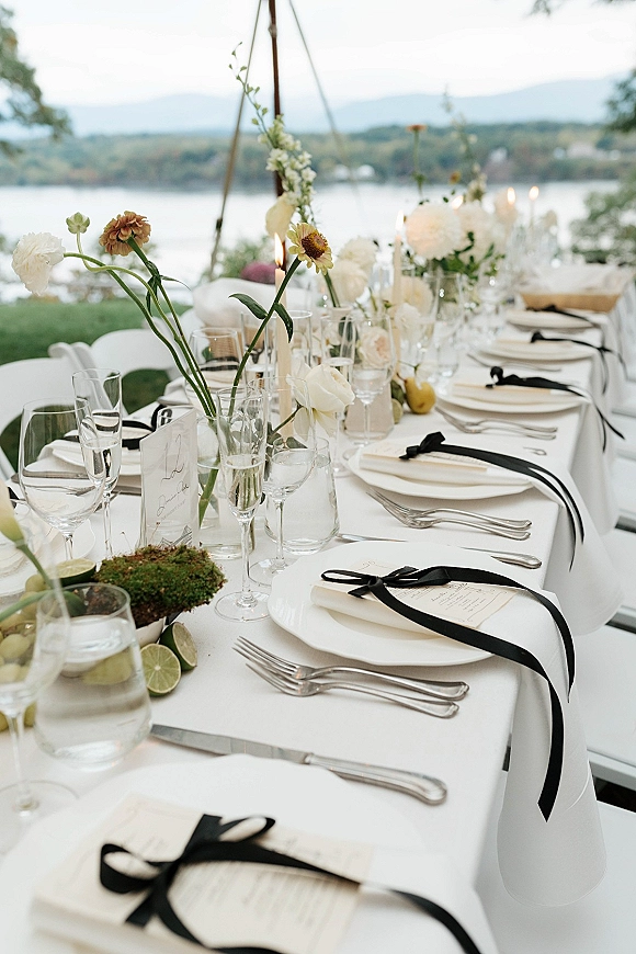 Reception tablescape on a long banquet table with white linens, black ribbon menus, taper candles, and white flowers overlooking a lakefront lawn