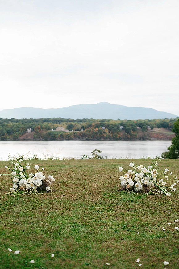 Ceremony aisle decor with grounded aisle flowers—low white blooms and greenery lining a grass lawn beside a river with mountains under clouds