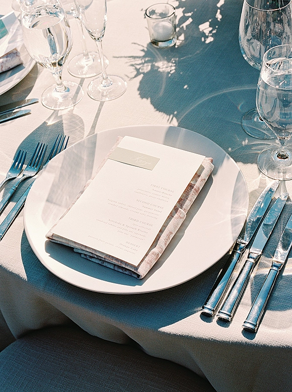 Wedding tablescape with a wedding place setting, white plate topped with menu card, linen napkin, place card, and silver flatware in sunlight