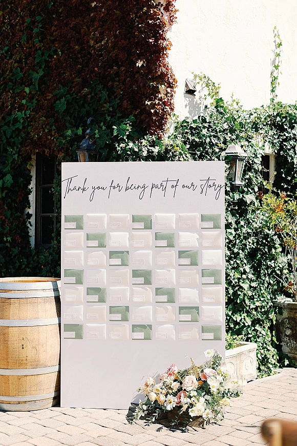 Wedding seating chart with escort card display, calligraphy sign and blush florals on a wooden barrel beside lanterns in an ivy courtyard