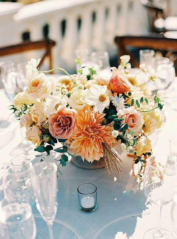 Reception centerpiece with a low floral centerpiece of roses, dahlias, daisies, and eucalyptus in a ceramic bowl beside a votive candle on an outdoor patio table