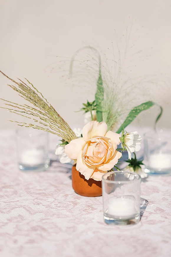 Wedding centerpiece with a peach rose in a terracotta bud vase, paired with glass votive candles on a patterned tablecloth setting
