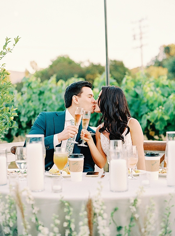 Wedding kiss as newlyweds raise champagne flutes at a candlelit sweetheart table, seated outdoors with vineyard greenery behind them