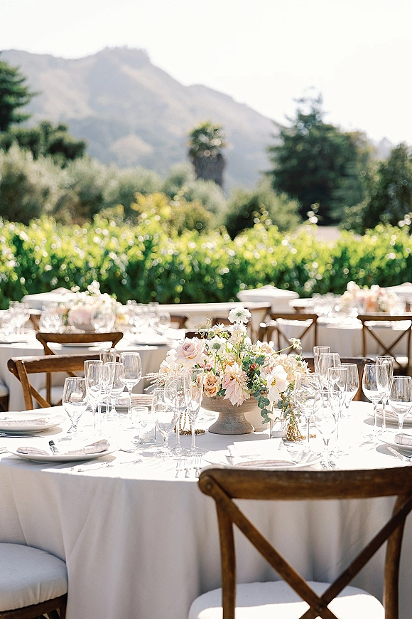 Reception tablescape on an outdoor wedding reception table with white linens, cross back chairs, floral compote centerpiece, and votive candles, mountain view beyond