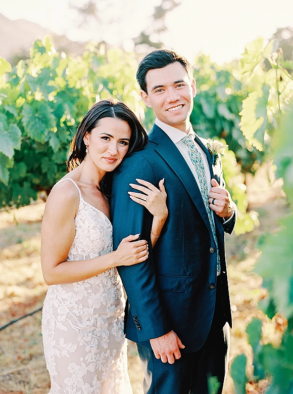 Couple portrait of bride hugging groom in a vineyard, her lace wedding dress and drop earrings catching warm golden hour light