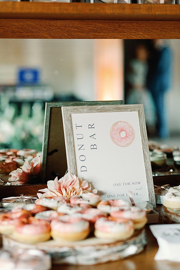 Donut bar wedding donut bar display with a framed sign, assorted donuts, pink flowers, and glass jars on a wooden table by a mirror backdrop