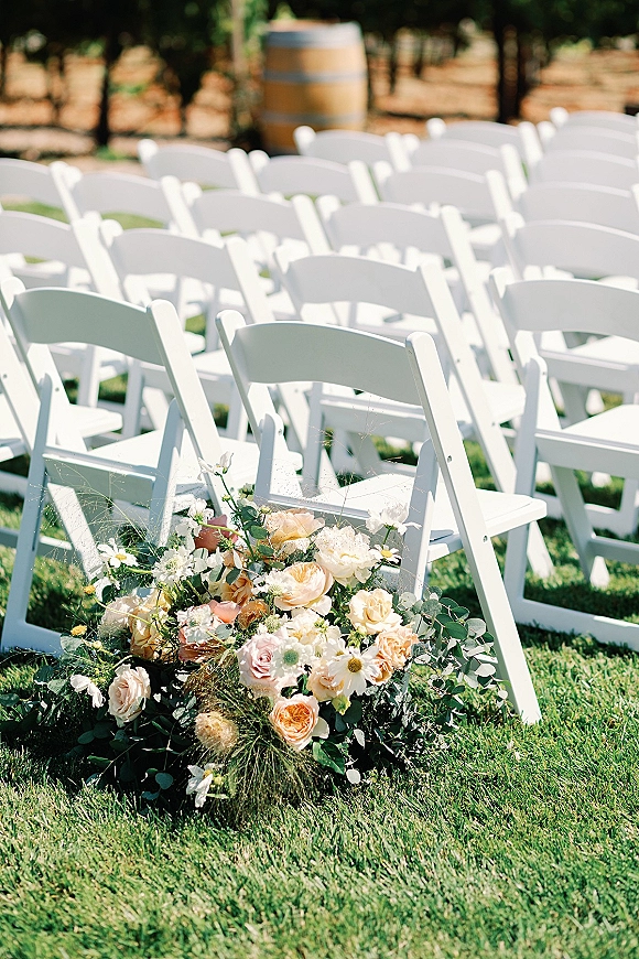 Ceremony seating with outdoor ceremony chairs in neat rows of white folding chairs, accented by rose and daisy florals on grass near trees