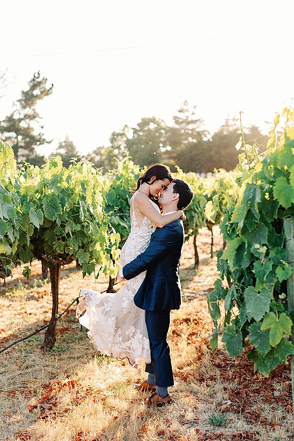 Wedding couple portrait of groom lifting bride in a lace wedding dress as they kiss between sunlit vineyard rows at golden hour