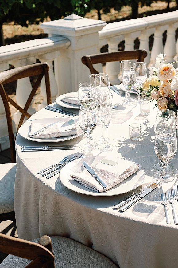 Reception tablescape with round wedding table setting, white tablecloth, silver flatware, candles, and floral centerpiece on a terrace with trees