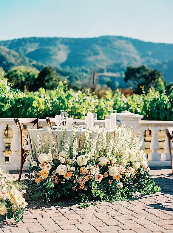 Sweetheart table decor with garden rose centerpiece, greenery garland and white taper candles on a stone terrace overlooking vineyard rows and mountains