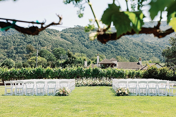 Ceremony setup with outdoor wedding ceremony seating, rows of white folding chairs and aisle florals on a lawn with vineyard and mountains beyond