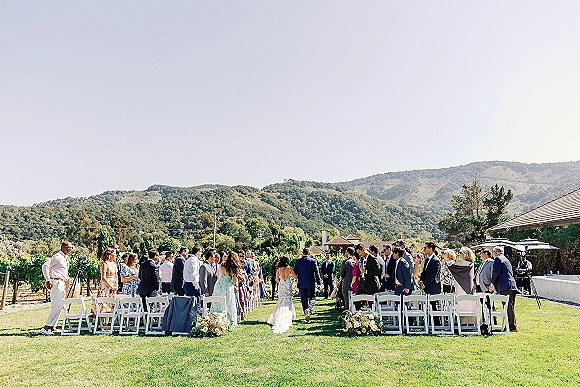 Outdoor ceremony wedding aisle walkway with guests in suits and dresses seated on white folding chairs on a vineyard lawn with mountain views